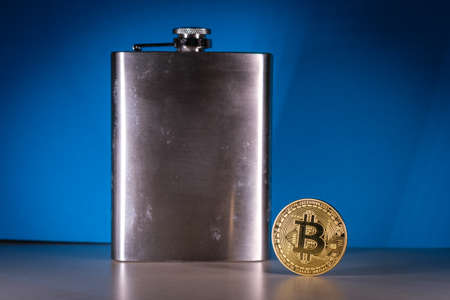 a bitcoin coin stands next to a steel hip flask