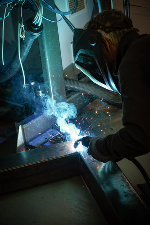 A person wearing a welding mask works on metal with a welding torch, creating sparks through the...