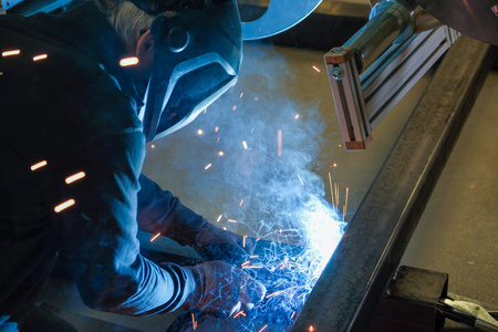 A person wearing a welding mask works on metal with a welding torch, creating sparks through the welding processの写真素材