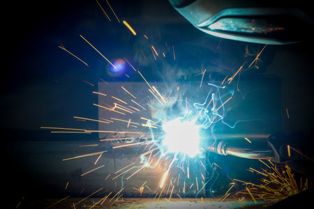 A person wearing a welding mask works on metal with a welding torch, creating sparks through the...