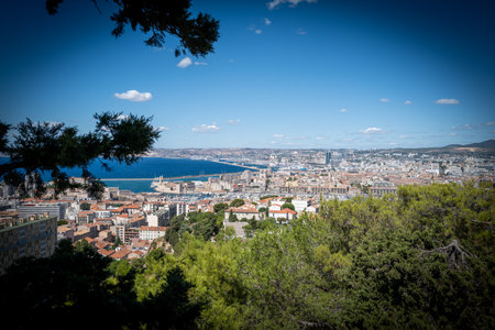 Panoramic view over Marseille, France, with the Old Port (Vieux-Port), Fort Saint-Nicolas, and the Mediterranean Sea under a clear blue sky. The photo captures the vibrant coastal cityscape with historical architecture, modern buildings, and the scenic waterfront. Tourists admire the stunning landscape from a viewpoint near Notre-Dame de la Garde, making this image ideal for travel, tourism, and Mediterranean lifestyle concepts.の写真素材