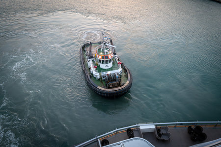 Close-up of a tugboat maneuvering in the harbor waters, assisting a large vessel during docking operations. Maritime transport scene with detailed view of the tugboat deck, safety equipment, and ocean waves in motion.の写真素材