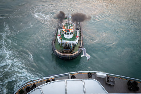 Close-up of a tugboat maneuvering in the harbor waters, assisting a large vessel during docking operations. Maritime transport scene with detailed view of the tugboat deck, safety equipment, and ocean waves in motion.の写真素材