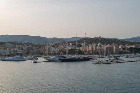 Panoramic view of Palma de Mallorca harbor with luxury yachts, sailboats, and modern city buildings against a backdrop of mountains and Bellver Castle. Scenic waterfront of Mallorca, Spain, at sunset with calm Mediterranean waters.の写真素材