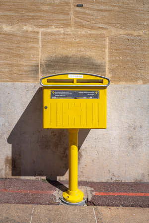 Bright yellow French mailbox fixed on a pole against a stone wall, photographed in sunlight. The vintage-style letterbox is commonly found in French towns and cities, symbolizing traditional mail services and communication. This urban detail captures European street culture and everyday life in France.の写真素材