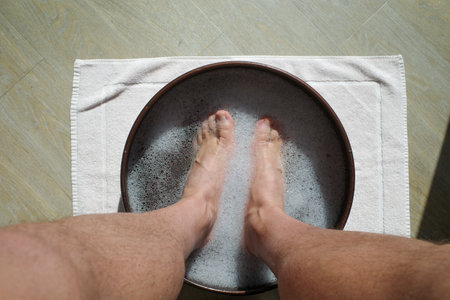 Close-up of male feet soaking in a wooden bowl filled with warm soapy water on a white towel, foot care and relaxation at home spa treatment. Concept of wellness, hygiene, pedicure, and stress relief therapy.の写真素材