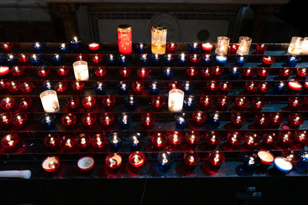 A peaceful scene of lit votive candles in red and blue glass holders inside a church or chapel. The glowing candlelight creates a warm and spiritual atmosphere, symbolizing prayer, remembrance, and faith. Some candles are housed in clear glass with religious icons, enhancing the sacred ambiance. Perfect for illustrating themes of spirituality, religion, meditation, mourning, and inner peace.の写真素材