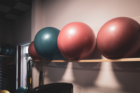 Row of colorful exercise balls neatly stored on a wooden wall rack inside a modern fitness studio. The arrangement features bright red and blue stability balls used for balance training, core workouts, physiotherapy, and rehabilitation exercises. The clean gym environment highlights essential functional training equipment and promotes an active, healthy lifestyle.の写真素材