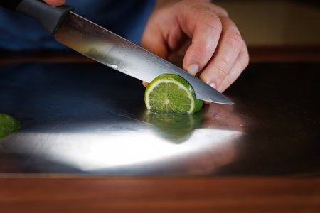Close-up of a person slicing a fresh lime with a sharp kitchen knife on a stainless-steel cutting board. The vibrant green citrus fruit is cut precisely, showing its juicy texture and natural pattern. This detailed food preparation scene highlights healthy cooking, fresh ingredients, kitchen skills, and culinary techniques in a home cooking environment.の写真素材