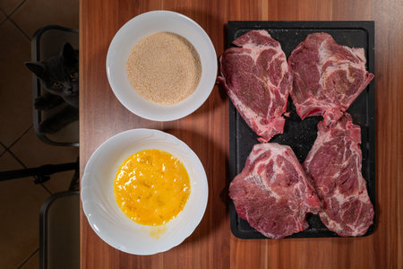 Overhead food photography showing the preparation of breaded meat with raw pork steaks on a black cutting board, a bowl of breadcrumbs, and a bowl of beaten eggs arranged on a wooden kitchen countertop. A curious gray cat peeks up from the side, adding a humorous and authentic home kitchen touch. The flat lay composition illustrates home cooking, schnitzel or cutlet preparation, breading process, and domestic lifestyle cooking. Ideal for food blogs, recipe content, cooking tutorials, and lifestyle photography with pets.の写真素材