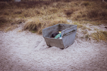 Environmental concept photograph showing an open metal trash container placed directly on a sandy beach surrounded by dry dune grass. Visible waste inside the container highlights issues of pollution, waste management, and human impact on fragile coastal ecosystems. The muted colors and natural setting create a realistic, documentary-style scene suitable for topics such as environmental protection, sustainability, coastal conservation, littering awareness, and climate responsibility.の写真素材