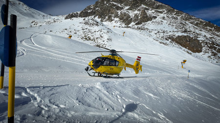 Yellow rescue helicopter parked on a snowy alpine ski slope surrounded by high mountain peaks under...