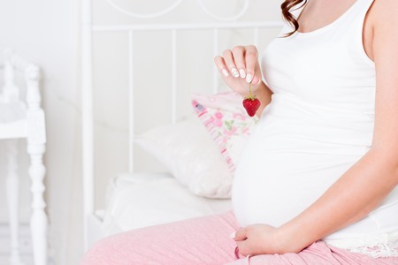 Happy pregnant woman sitting on the bed. A young pregnant woman is sitting in the bedroom. Pregnant woman relaxing. Young pregnant woman eating strawberries.の写真素材