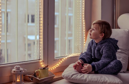 Little cute boy sitting in the window. Christmas mood. Holidays. A cozy evening. Mug of hot cocoa with marshmelow. Winter eveninの写真素材