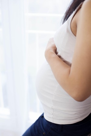 Beautiful young Asian pregnant woman standing by the window and touching her tummy. Pregnancy. Health. Motherhood. A cozy portrait of a pregnant woman.の写真素材