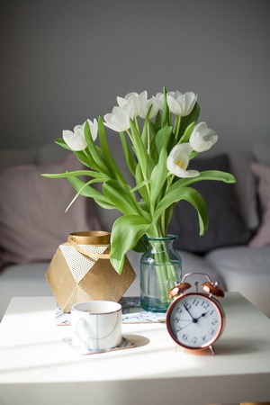 Interior. Room. A bouquet of tulips in a glass vase, a gold candlestick, a mug of hot coffee, an alarm clock on a white wooden table. Sofa, pillows. Spring. It's cozy.の写真素材