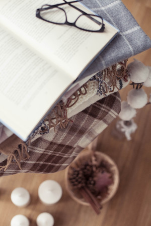 Plaids. Autumn cozy interior. On a wooden chair is a stack of warm blankets. Candles, leaves, cones, basket, cinnamon. Book and glasses. Autumn. Winter.の写真素材