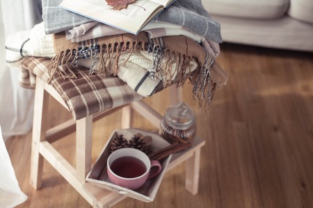 Plaids. A cup of hot tea. Autumn cozy interior. On a wooden chair is a stack of warm blankets. Candles, leaves, cones, basket, cinnamon. Book and glasses. Autumn. Winter.の写真素材