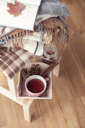 Plaids. A cup of hot tea. Autumn cozy interior. On a wooden chair is a stack of warm blankets. Candles, leaves, cones, basket, cinnamon. Book and glasses. Autumn. Winter.の写真素材