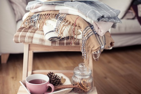 Plaids. A cup of hot tea. Autumn cozy interior. On a wooden chair is a stack of warm blankets. Candles, leaves, cones, basket, cinnamon. Book and glasses. Autumn. Winter.の写真素材