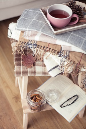 Plaids. A cup of hot tea. Autumn cozy interior. On a wooden chair is a stack of warm blankets. Candles, leaves, cones, basket, cinnamon. Book and glasses. Autumn.の写真素材