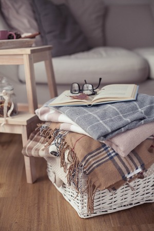 Plaids. A cup of hot tea. Autumn cozy interior. On a wooden chair is a stack of warm blankets. Candles, leaves, cones, basket, cinnamon. Book and glasses. Autumn.の写真素材