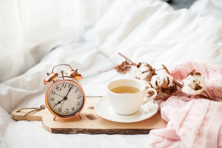 Alarm clock, a beautiful white cup with tea on the bed, pink knitted blanket. Breakfast in bed....