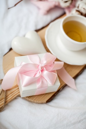 White mug with tea, gift box with ribbon on the bed. Breakfast in bed. Cozy. Pink plaid. Cotton. Spring.の写真素材