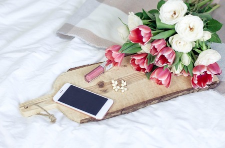 A wooden board, a plaid, a bouquet of tulips, decorations, a telephone, cosmetics lie on a white bed. Spring.の写真素材