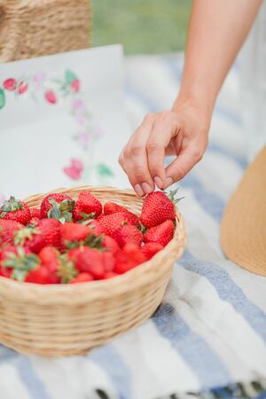 Close-up. Young beautiful pregnant woman in white dress is sitting in the meadow and eating strawberries. Picnic. Motherhood.の写真素材