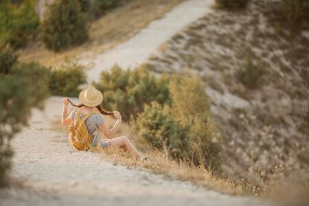 Beautiful red-haired young woman in a hat with a backpack sits on a cliff in the mountains.の写真素材