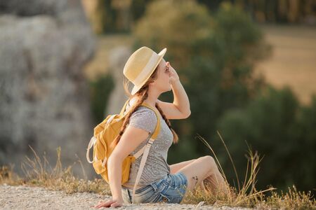 Beautiful red-haired young woman in a hat with a backpack sits on a cliff in the mountains.の写真素材