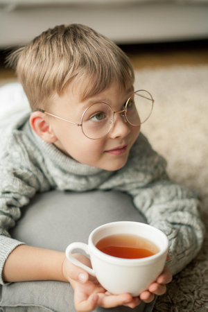 A beautiful little boy in a knitted sweater is reading by the window with a mug of hot tea. Cozy. Autumn. Fall mood.の写真素材