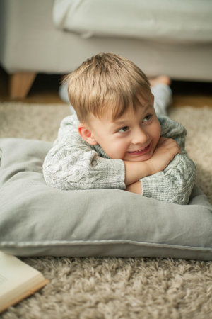 A beautiful little boy in a knitted sweater is reading by the window with a mug of hot tea. Cozy. Autumn. Fall mood.の写真素材