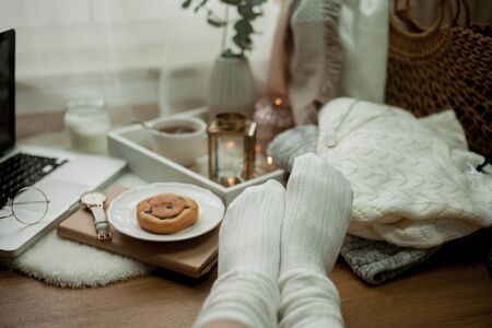 Toned photo. Autumn decor. Women's legs in knitted socks, a laptop, a mug of hot tea, a bun, candles. Cozy. Autumn. Fall mood.の写真素材