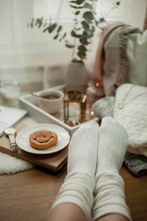 Toned photo. Autumn decor. Women's legs in knitted socks, a laptop, a mug of hot tea, a bun,...