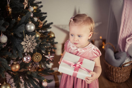 Beautiful little girl with blue eyes in a pink dress holds a gift near the Christmas tree. Christmas. New Year. Holidays.の写真素材