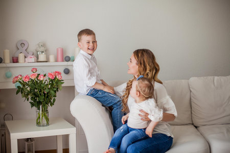Beautiful young mother, little son, little daughter are sitting on the sofa at home. Mothers Day. A happy family.の写真素材