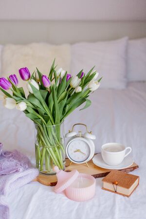 A bouquet of tulips in a glass vase, a white alarm clock, a candle, a note, a cappuccino mug on a wooden board are standing on the bed. Womens things. Breakfast in bed.の写真素材