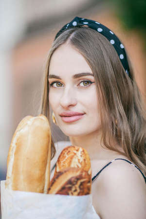Beautiful young woman in a european city holds a french baguette. French style. City.の写真素材