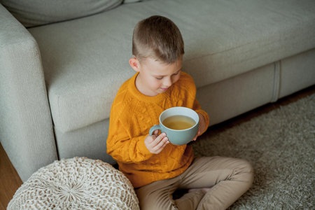 A little cute boy in an orange sweater is drinking tea. Cozy portrait of a boy sitting at home.の写真素材