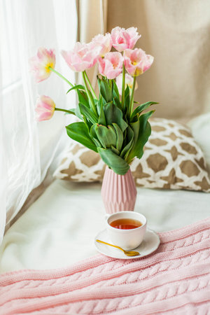 A mug of hot tea, a pink knitted blanket, a bouquet of tulips in a vase. Breakfast in bed. Cozy.の写真素材
