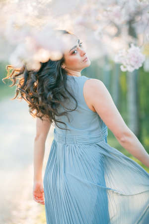 Beautiful young woman with curly hair drinks pink champagne in the blooming sakura garden. Picnic in nature. Spring.の写真素材
