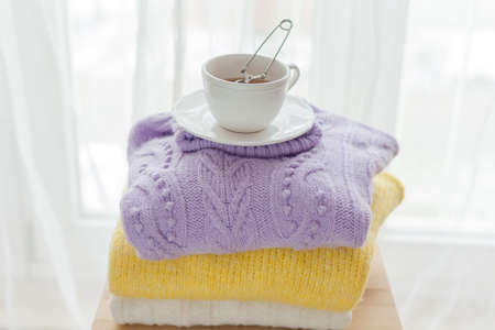 A stack of spring knitted clothes on a wooden chair, a mug of hot tea with tea leaves. Yellow, Purple colors.の写真素材