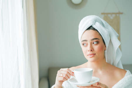 After a shower, a young woman in a bathrobe with a towel on her head drinks coffee and looks out the window.の写真素材