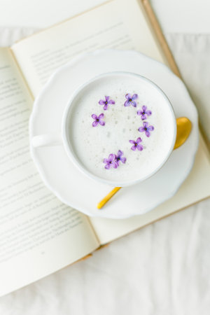 Mug of cappuccino on a beige background. Lilac flowers, book. Rest at home. Quarantine. Cozy.の写真素材