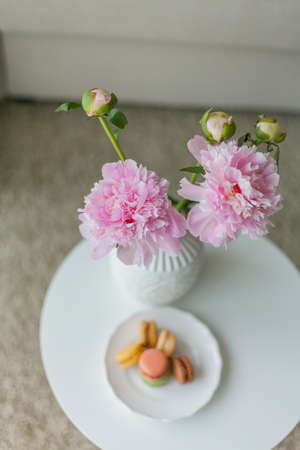 Living room interior. Bouquet of pink peonies in a vase, multicolored French macaroon pastries. Cozy.の写真素材