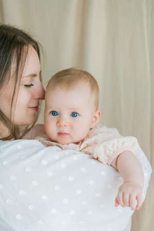 A young beautiful mother in a white dress is holding her little blue-eyed daughter of 5 months in her arms.の写真素材