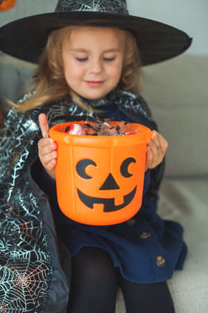 Little cute girl 3 years old in a witch costume with a pumpkin bucket full of sweets celebrates Halloween at home with decorations. Holidays.の写真素材