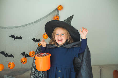 Little cute girl 3 years old in a witch costume with a pumpkin bucket full of sweets celebrates Halloween at home with decorations. Holidays.の写真素材